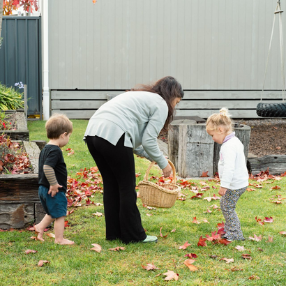 Woman and two children in backyard collecting fallen leaves with a basket.