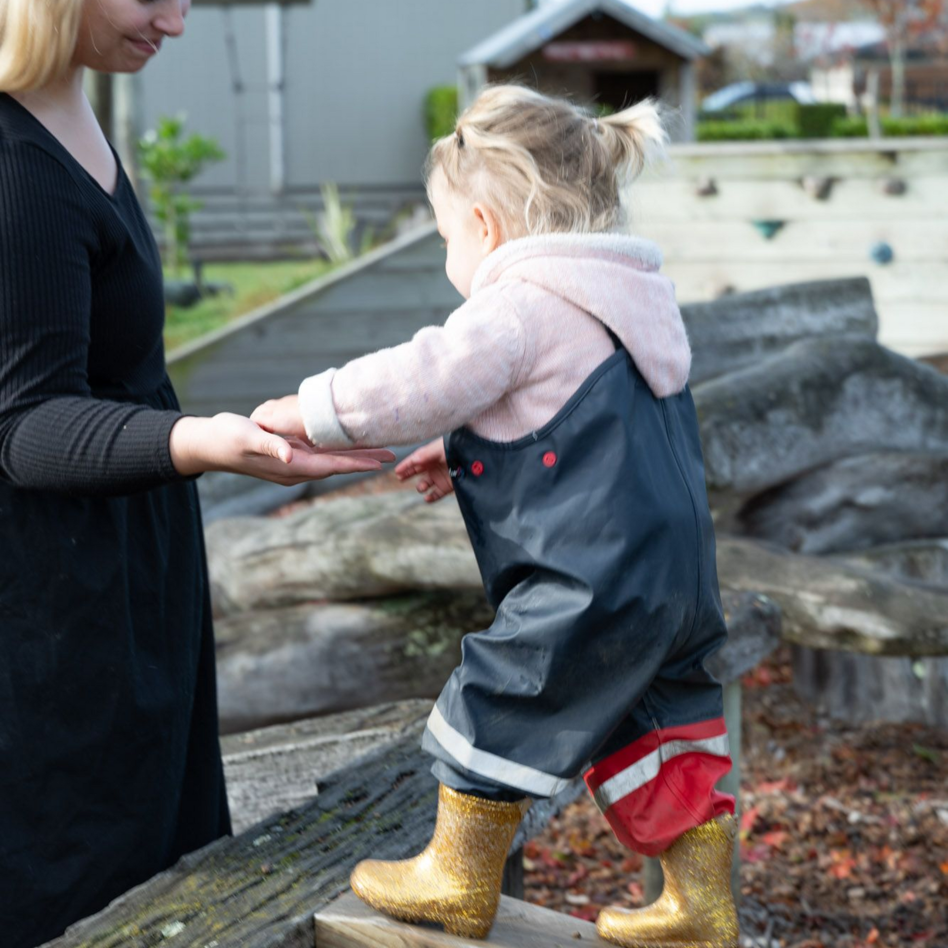 Woman helping a toddler with blond pigtails wearing gold boots, navy overalls, on a wood structure.
