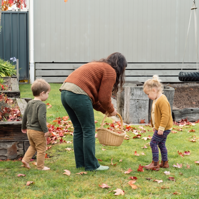 Woman and two children in backyard collecting fallen leaves with a basket.
