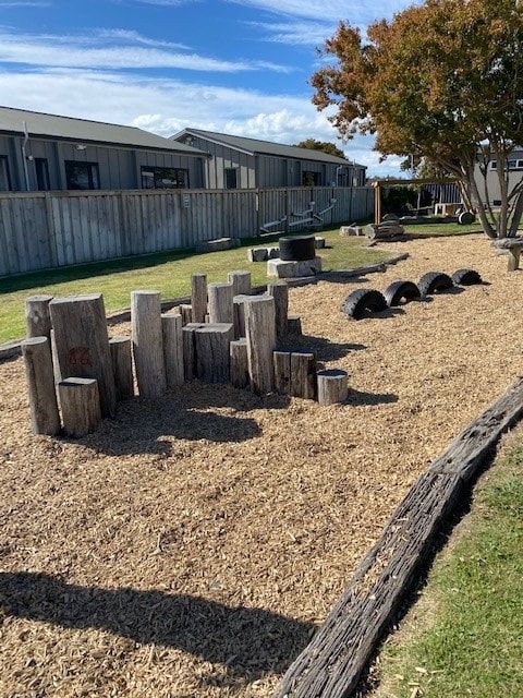 Playground with wood blocks, tires, and wood chip ground cover. Buildings and fence in the background.