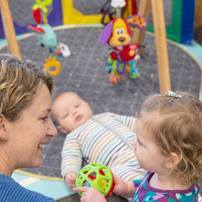 Woman and toddler looking at a toy, with sleeping baby on a playmat. Toys are overhead.