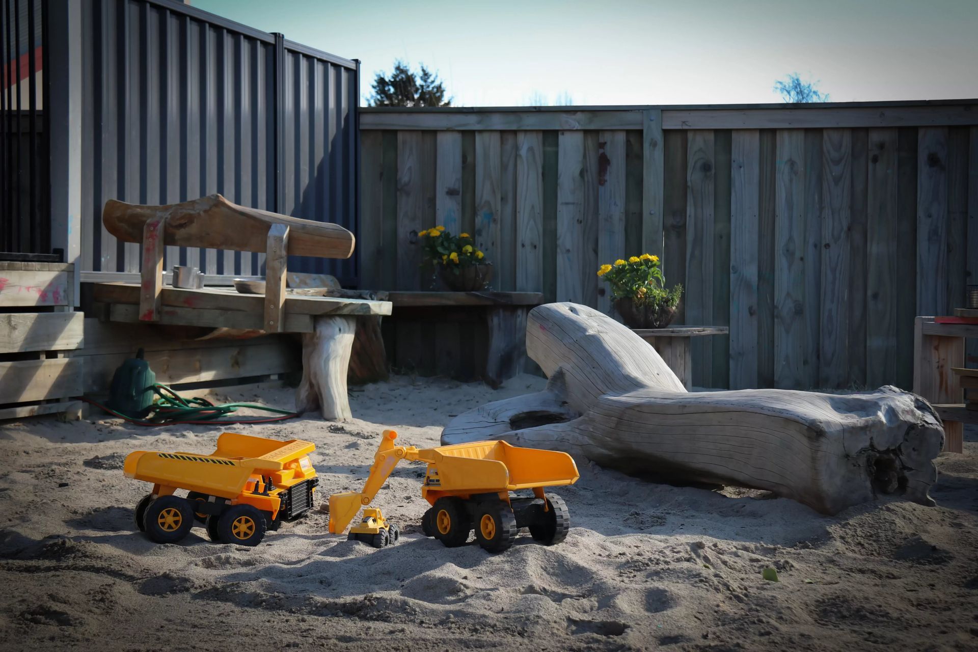 Sandbox with yellow toy trucks, wooden bench, and fence.