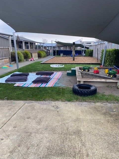 Playground area with sandpit, shaded lawn, and building in background.