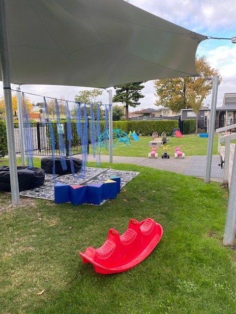 Playground with red teeter-totter on grass, shaded by canopy. Children play in the background.