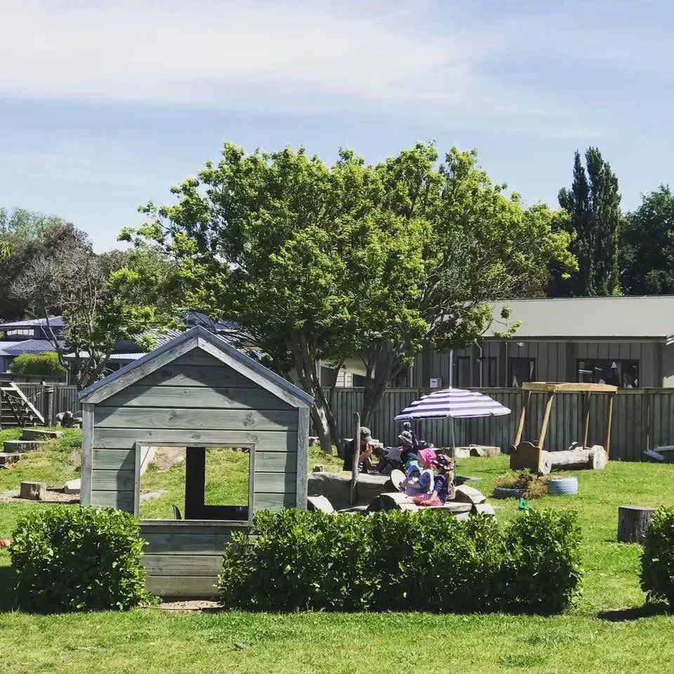 Children playing outdoors at a daycare. Features a playhouse, parasol, and wooden swing set on a sunny day.