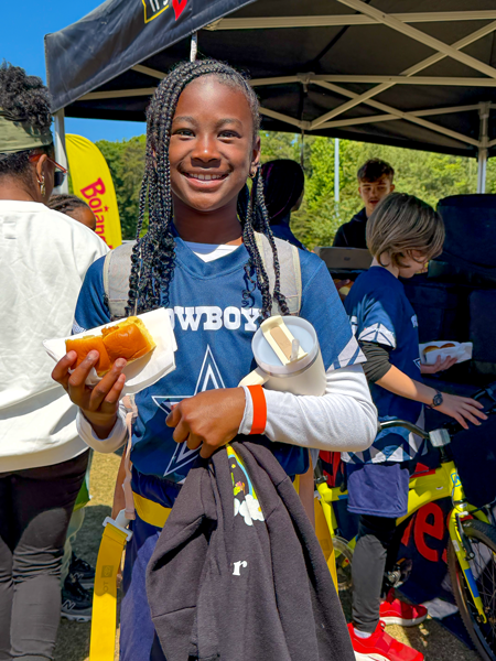 Smiling flag football player in blue jersey holding a snack from Bojangles at an outdoor event booth under a canopy.