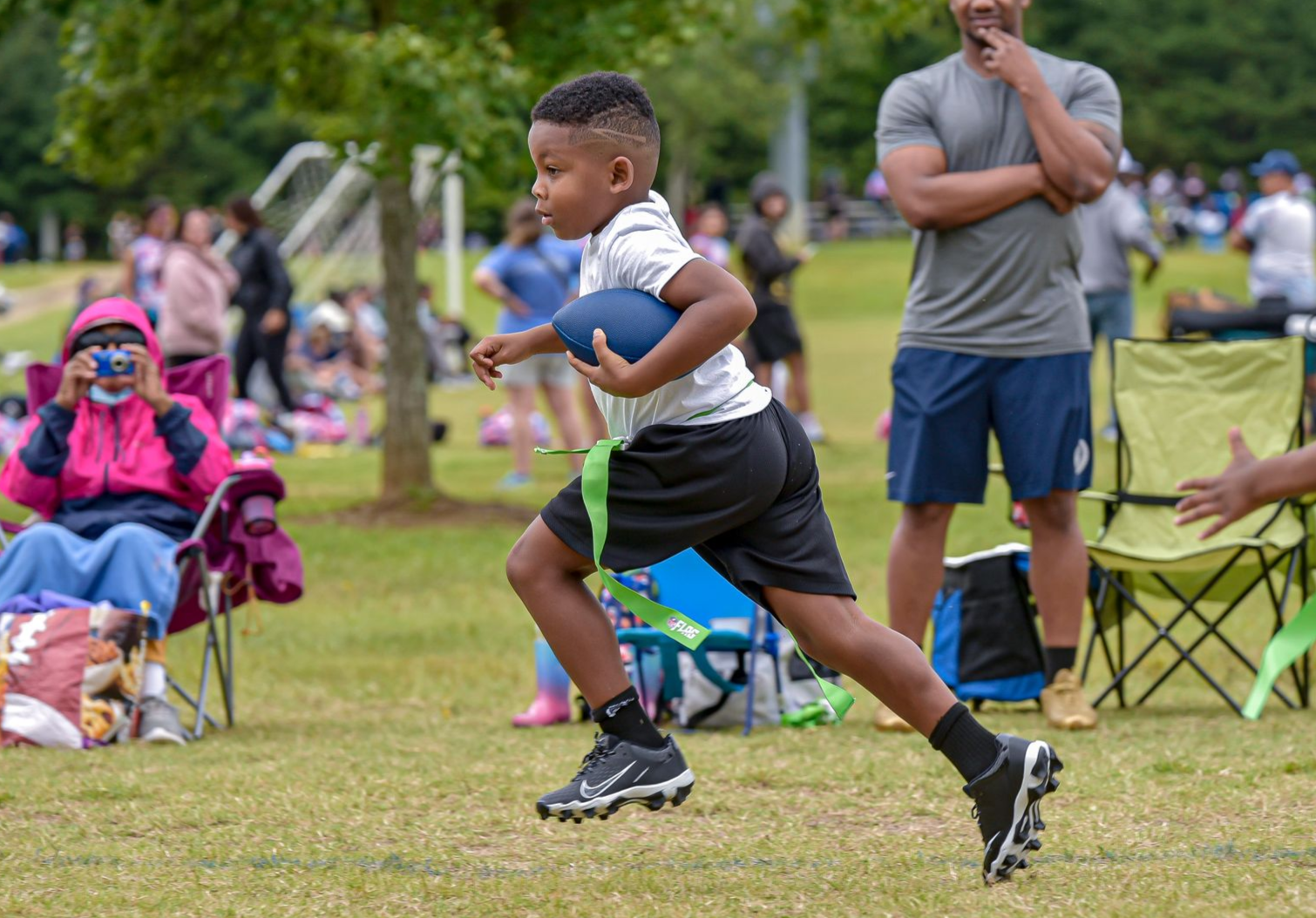 Kids running in a grassy park while spectators watch from folding chairs.