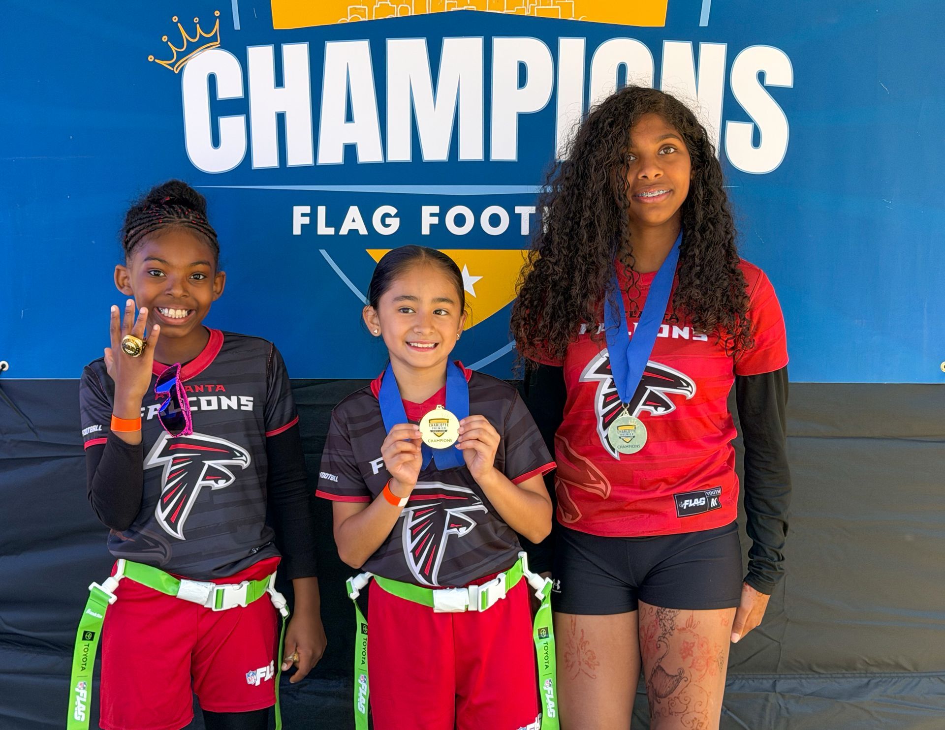 Three flag football champions holding medals and trophies in front of a CHAMPIONS banner