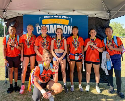 Group of flag football players in orange jerseys posing under a tent in front of a 