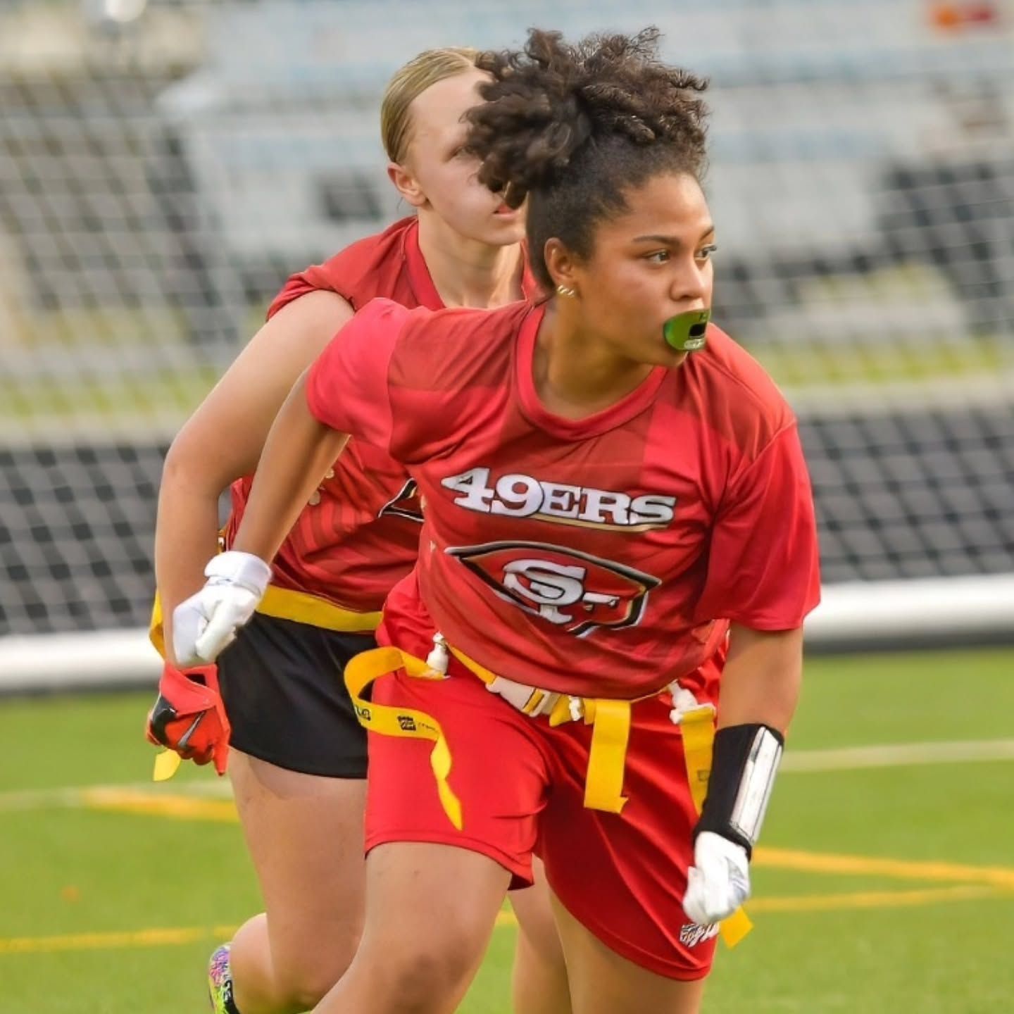 Youth flag football player in red and yellow runs with the ball on a grassy field, teammates and tents behind
