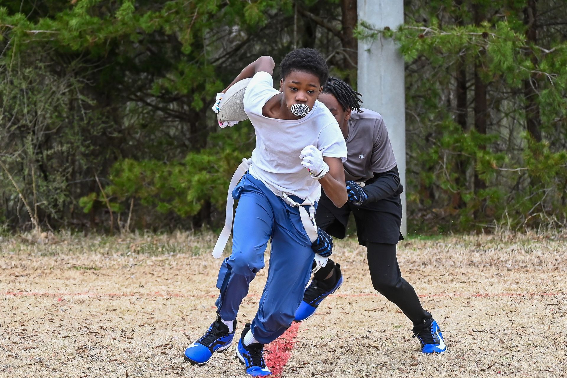 Two children playing flag football on a grassy field, one in a white shirt and blue pants in the foreground.