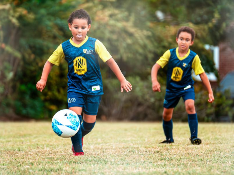 Two children in blue soccer uniforms play on a grassy field, one dribbling a ball.