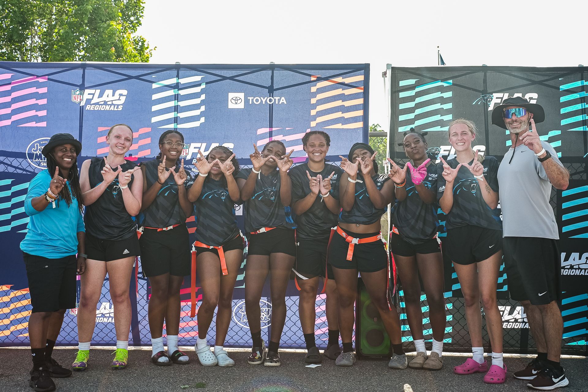 Eight flag football players posing on a finish line podium, holding medals and fists up in front of sponsor banners.