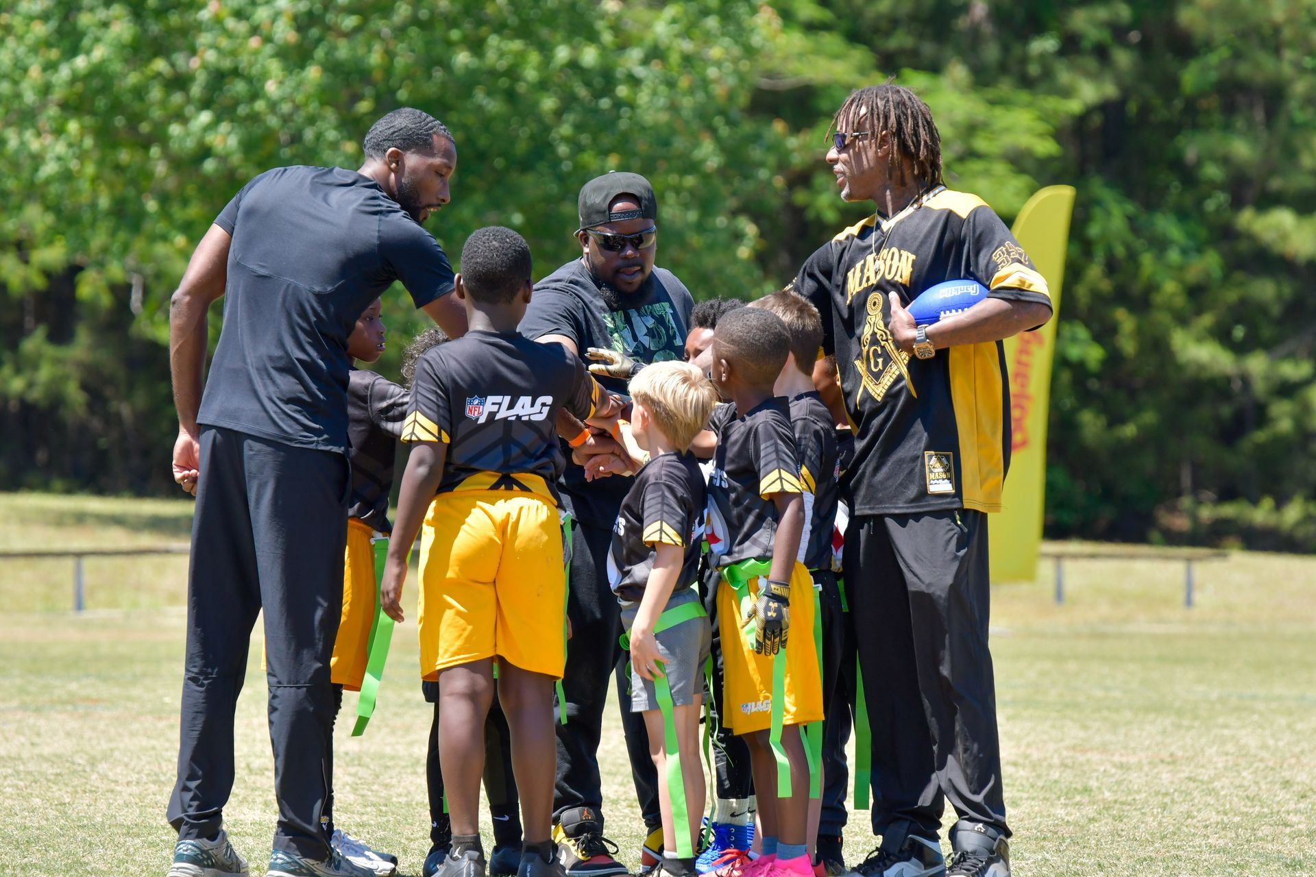 Youth flag football team huddles with coaches on a grassy field, preparing for practice.
