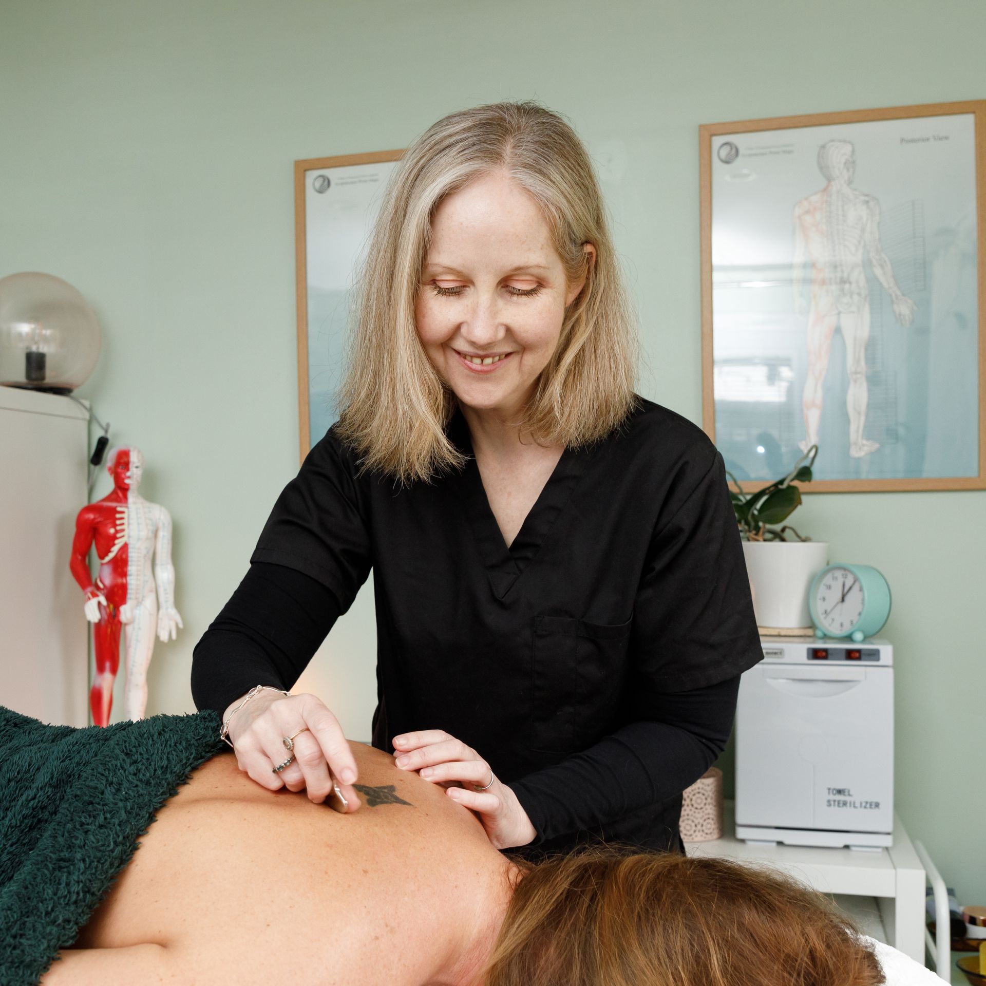 Woman in black scrubs performing back treatment. Beige-walled room, anatomical posters.