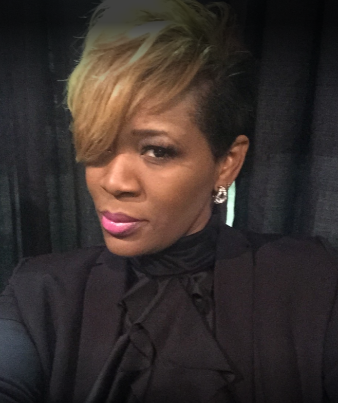 Woman with blonde-tipped hair and pink lipstick wears a black jacket and gazes at the camera.