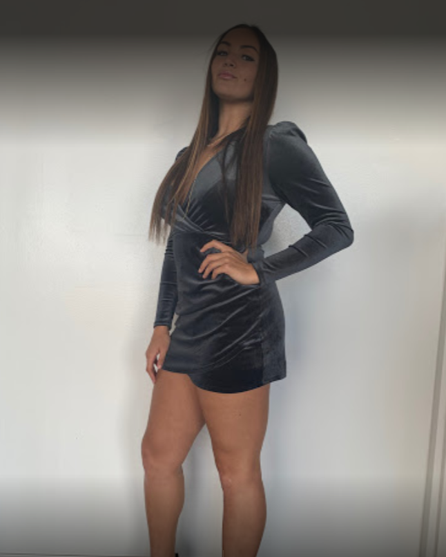 Woman in a gray velvet dress poses with hand on hip against a plain white wall.