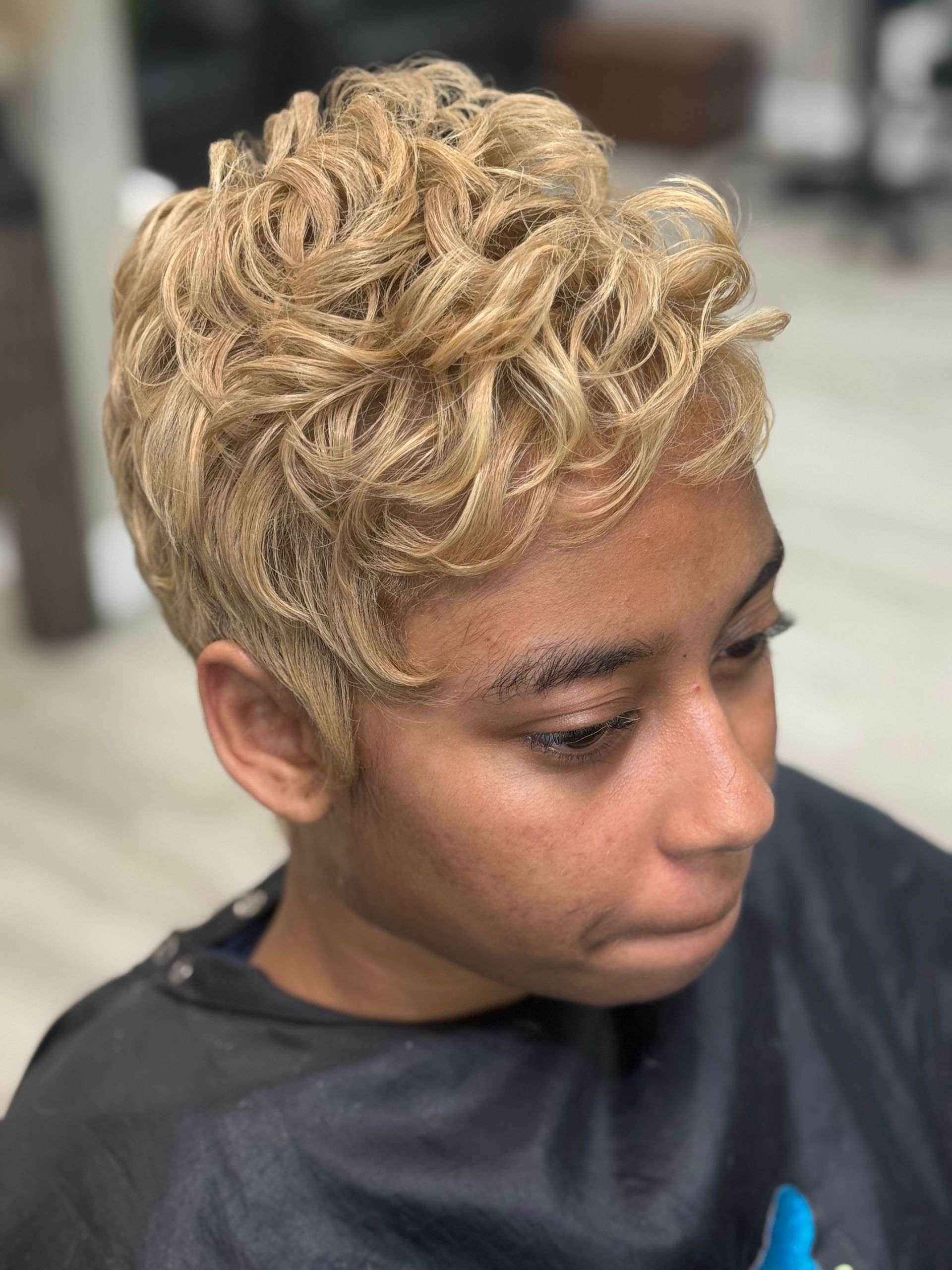 Person with short, curly blonde hair, looking down. Wearing a black salon cape.