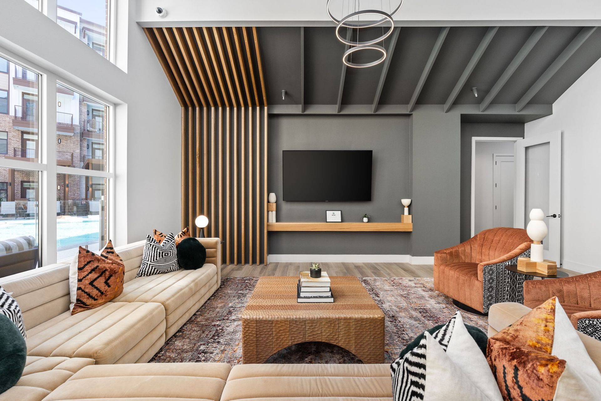 A modern living room with a large tan sectional, a wooden coffee table, orange accent chairs, and a slatted wood feature.
