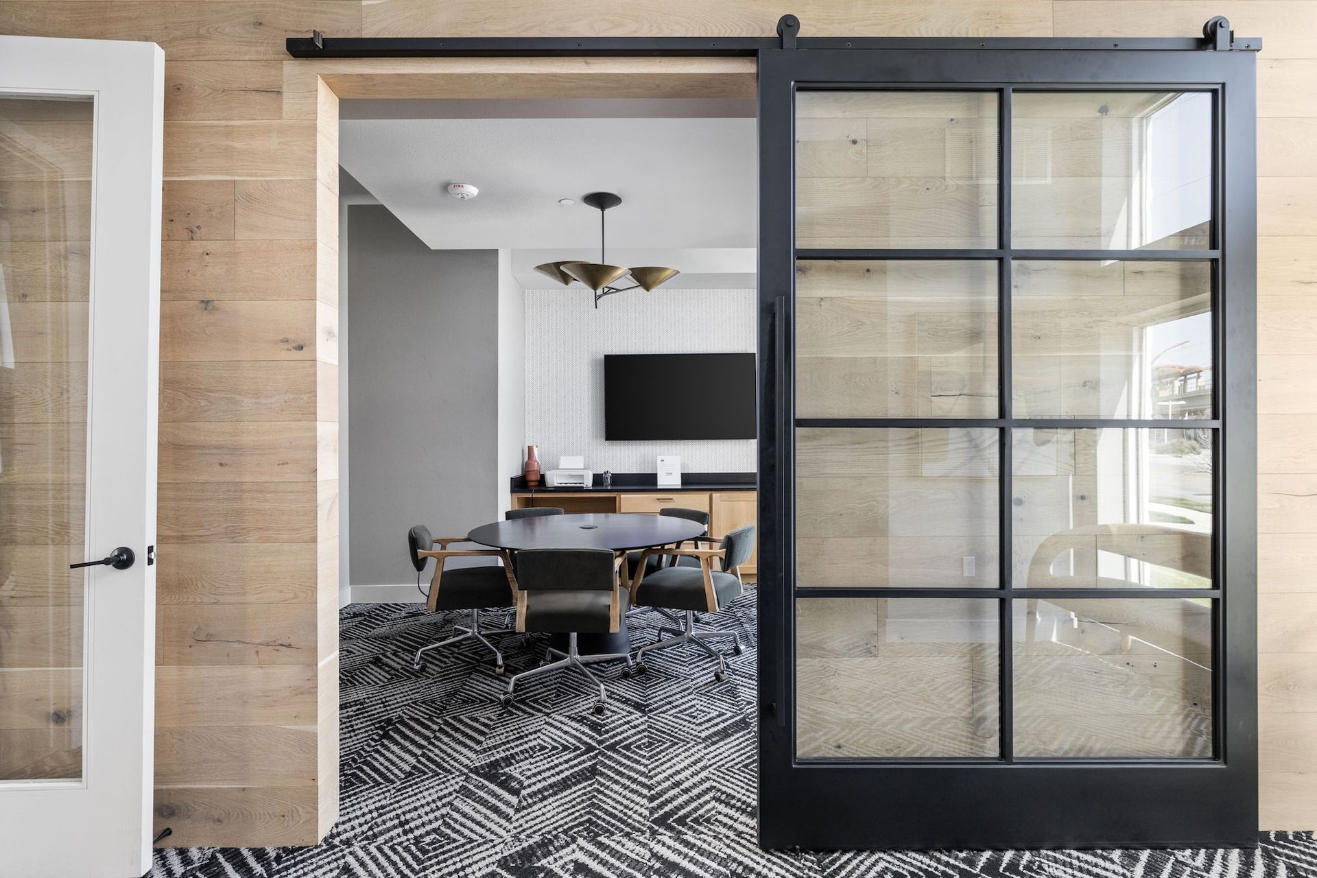 Black sliding glass barn door leading to an office space with a round conference table, chairs, and a wall-mounted TV.