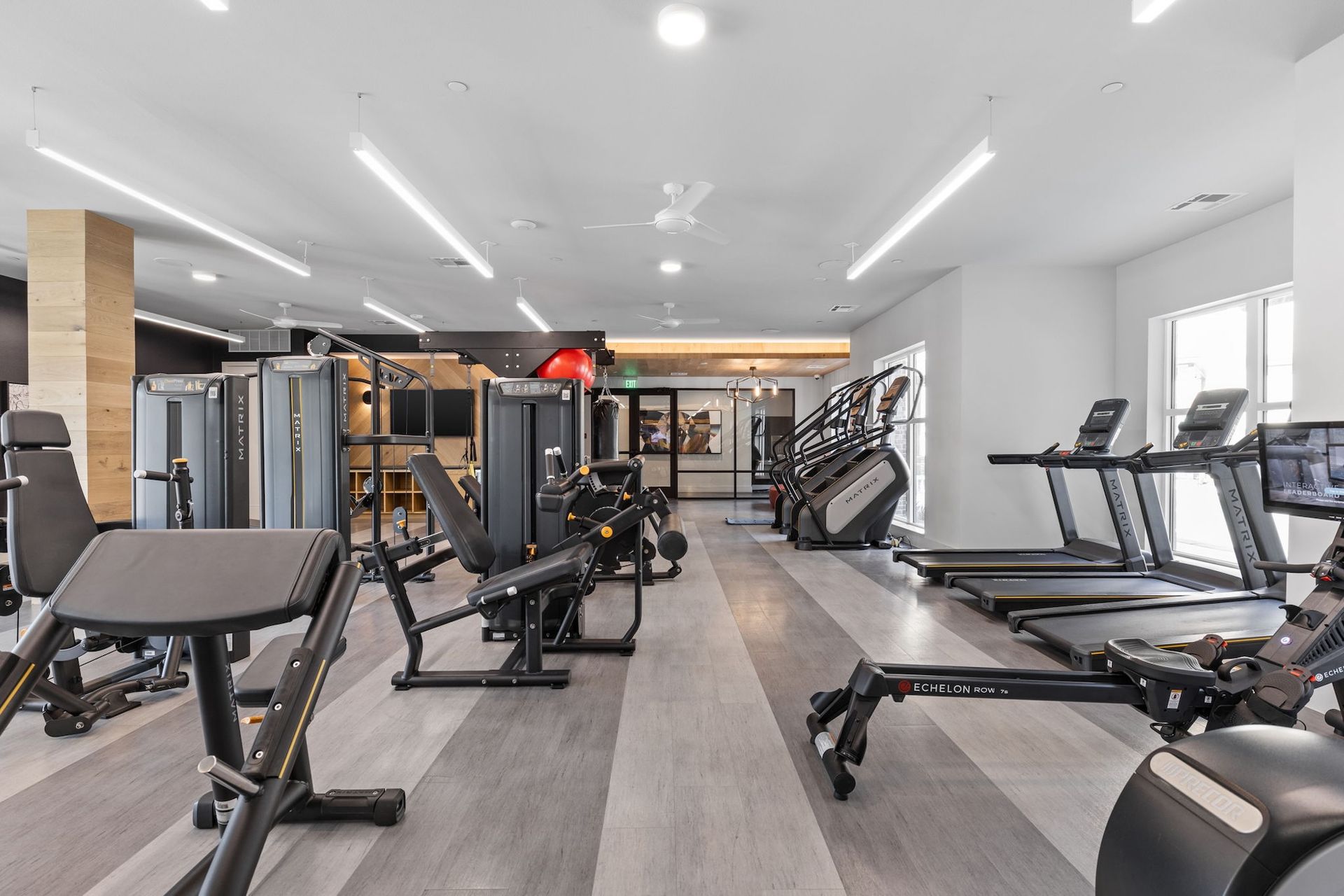 A modern fitness center featuring strength training machines, treadmills, and a rowing machine on grey wood flooring.
