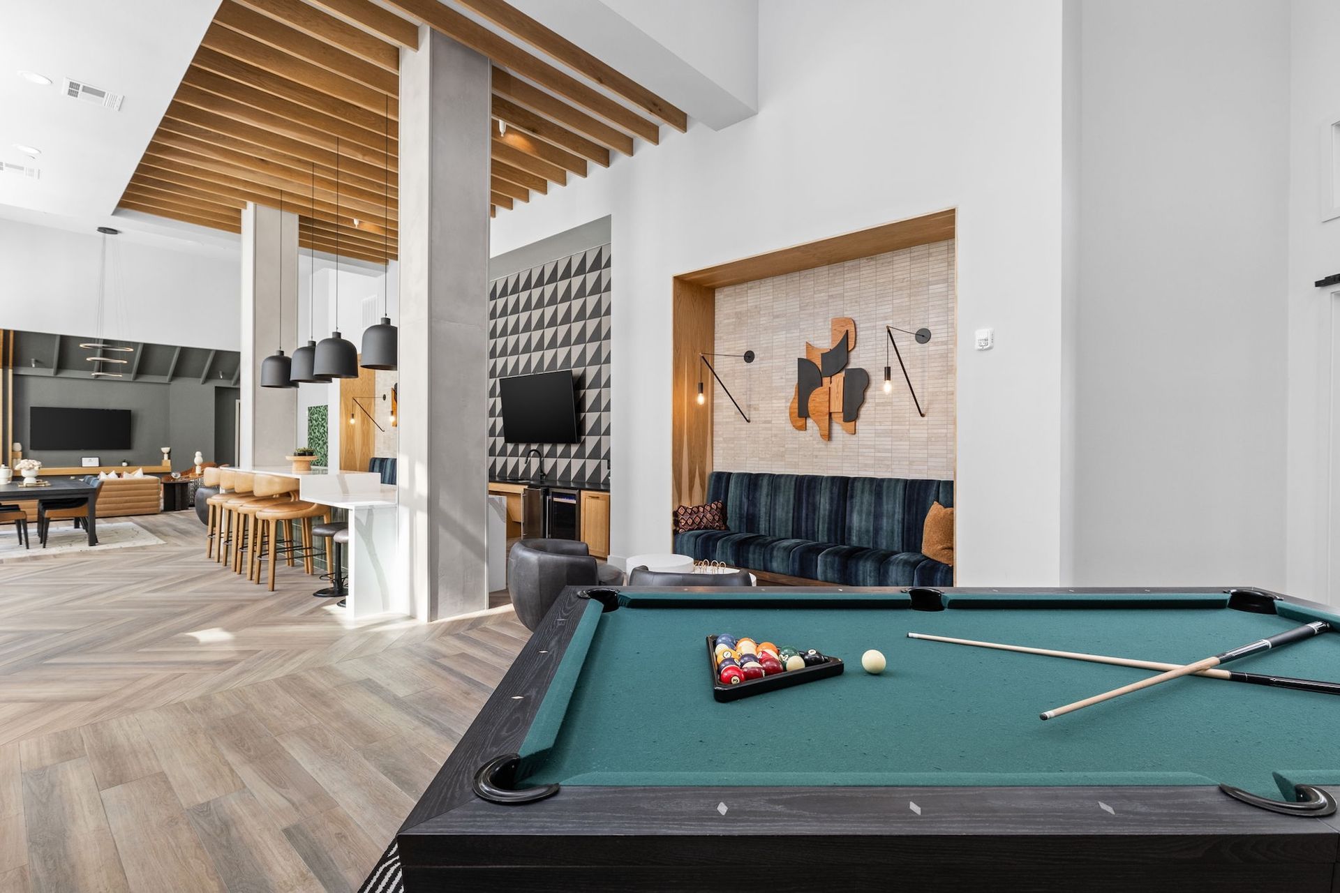 A modern common room with a billiards table in the foreground and a seating area, bar, and TVs in the background.