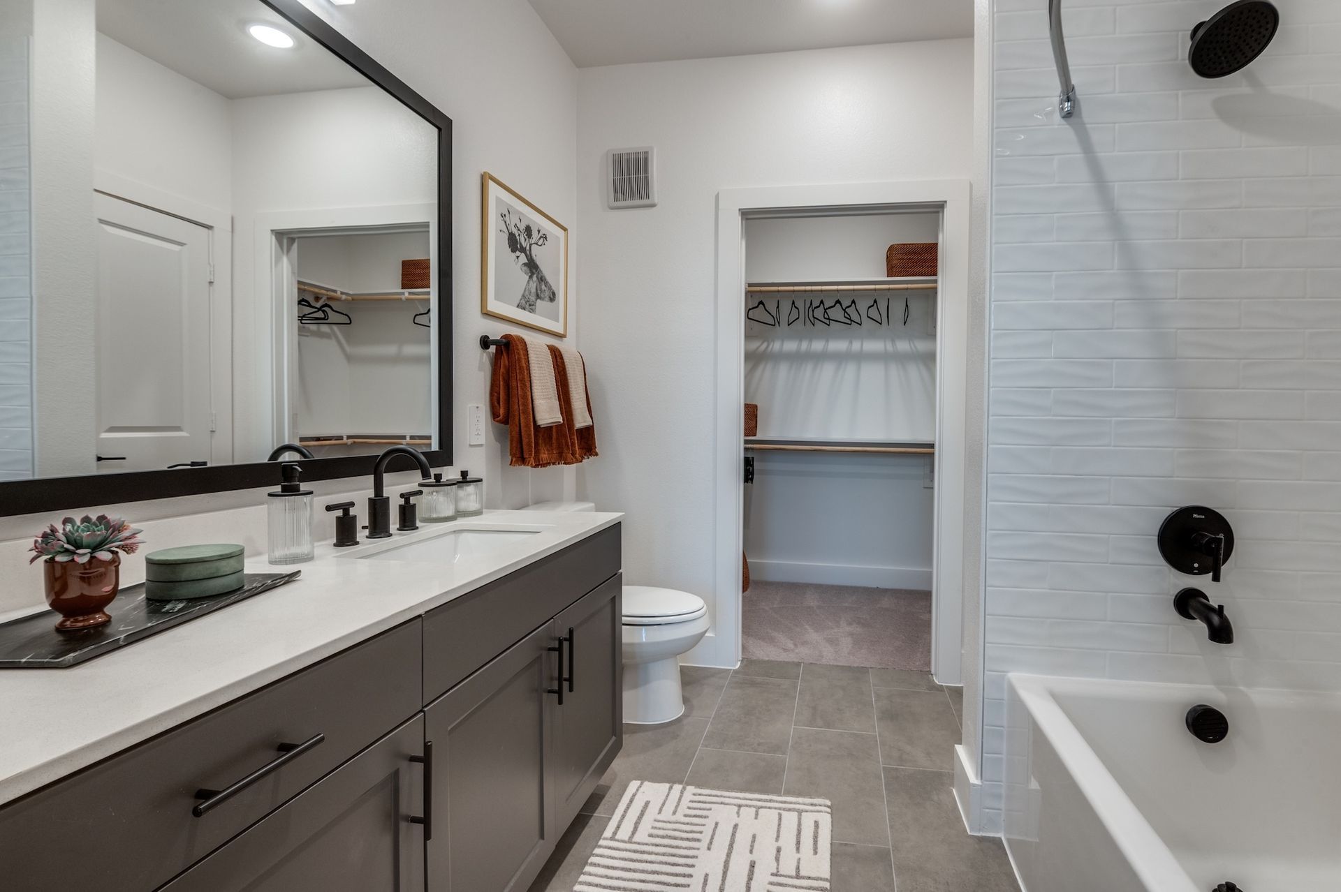 A modern bathroom with a grey vanity, white countertops, a large mirror, a tiled shower, and an adjacent walk-in closet.