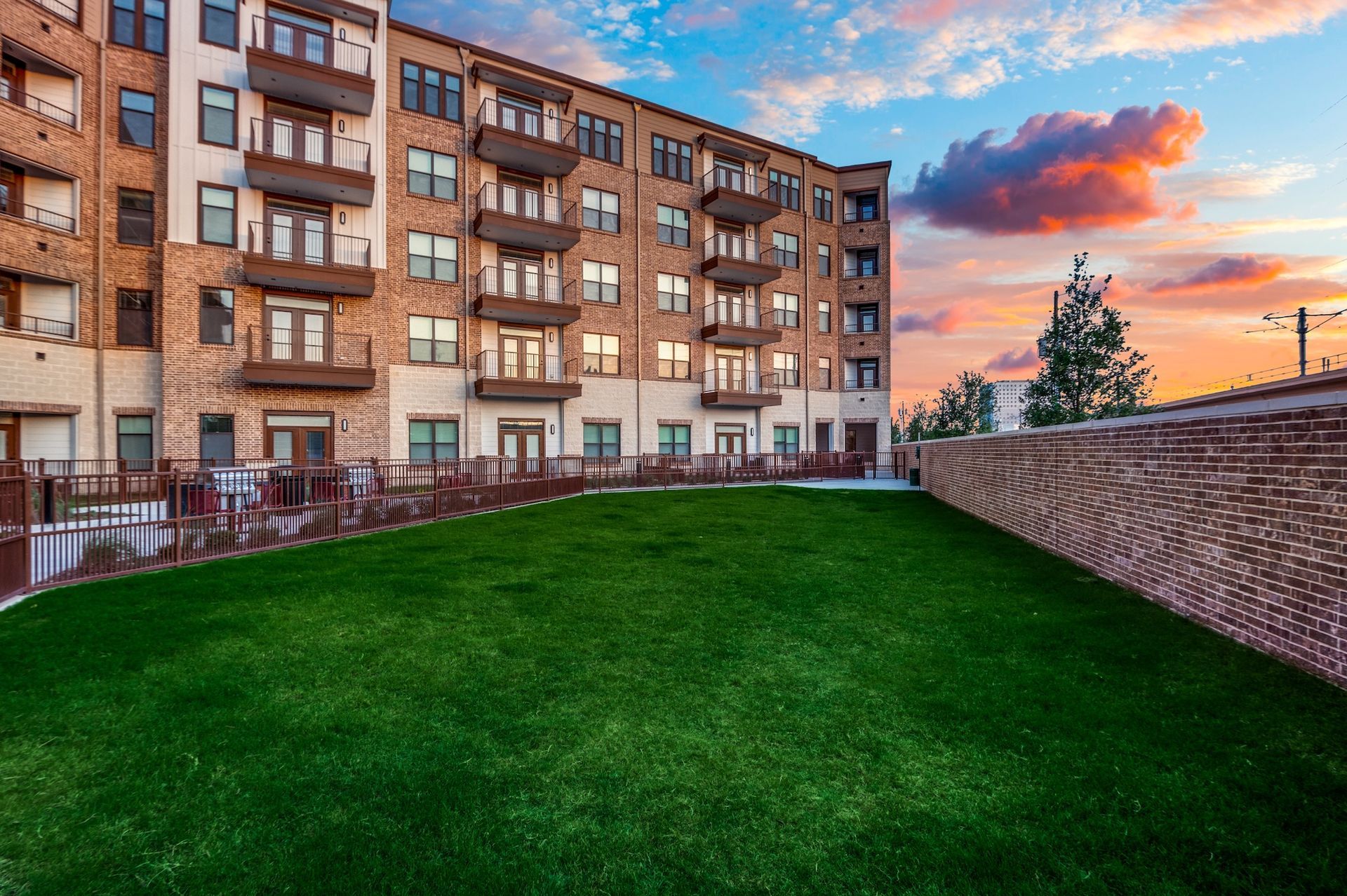 Exterior view of an apartment building with balconies, set against a green lawn and a sunset sky.