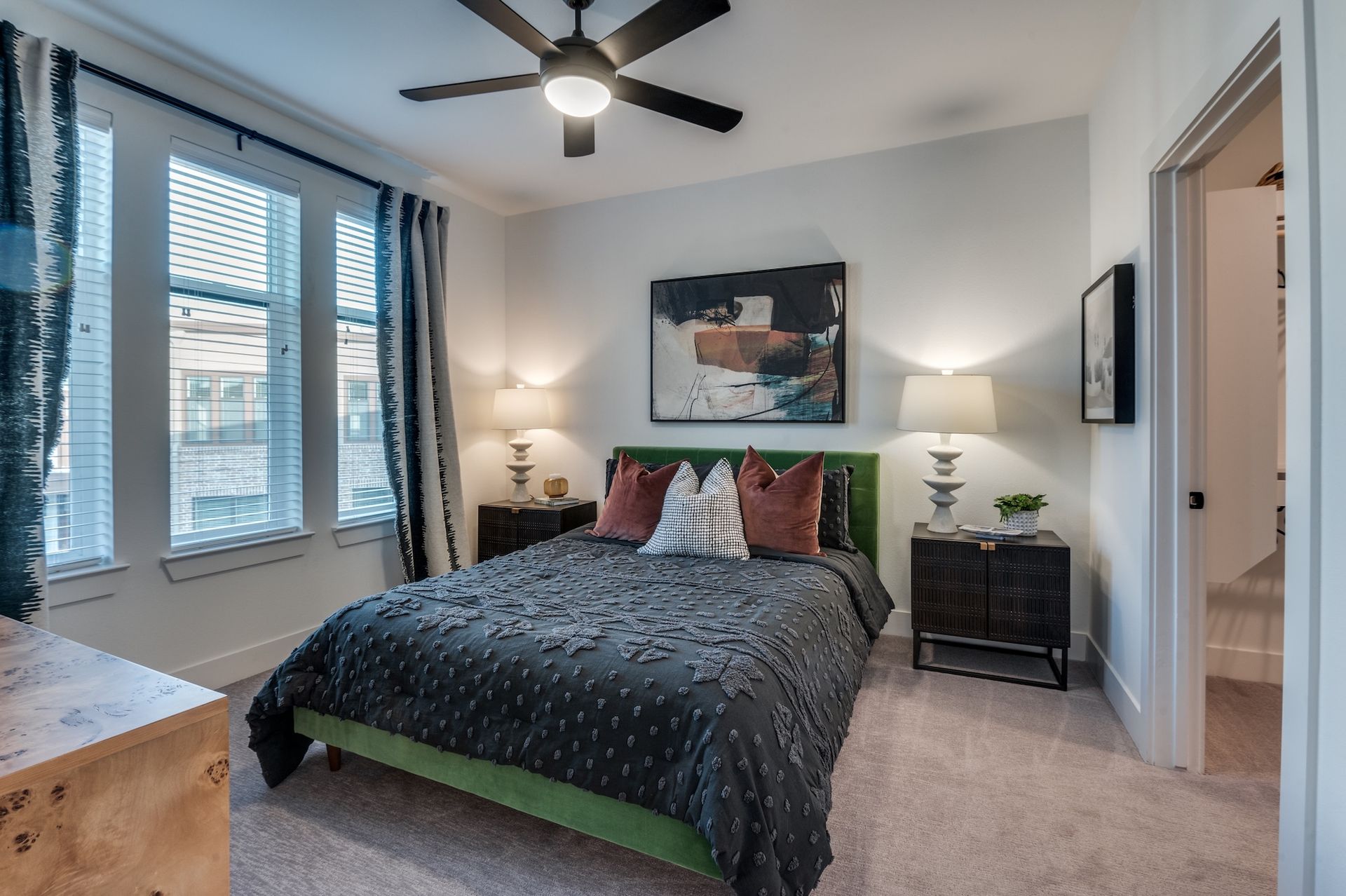 A modern bedroom featuring a bed with a green frame, patterned dark bedding, two nightstands with lamps, and large windows.