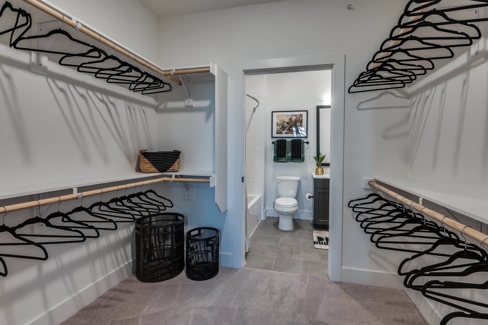 A walk-in closet with shelves and rows of black hangers on both sides, leading to a small bathroom with a toilet.