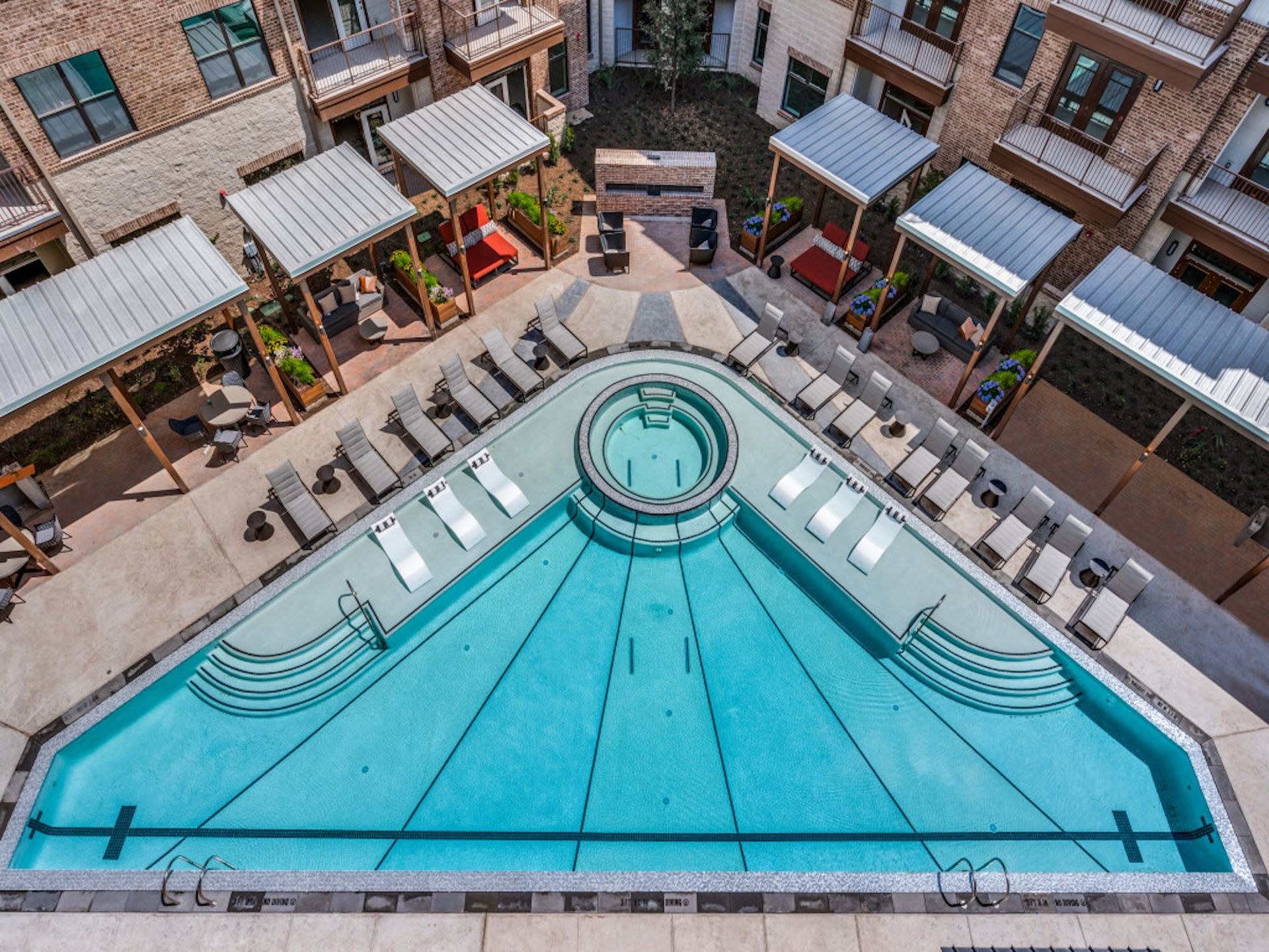 High-angle view of a turquoise, triangle-shaped apartment pool surrounded by sun loungers and shaded cabanas.