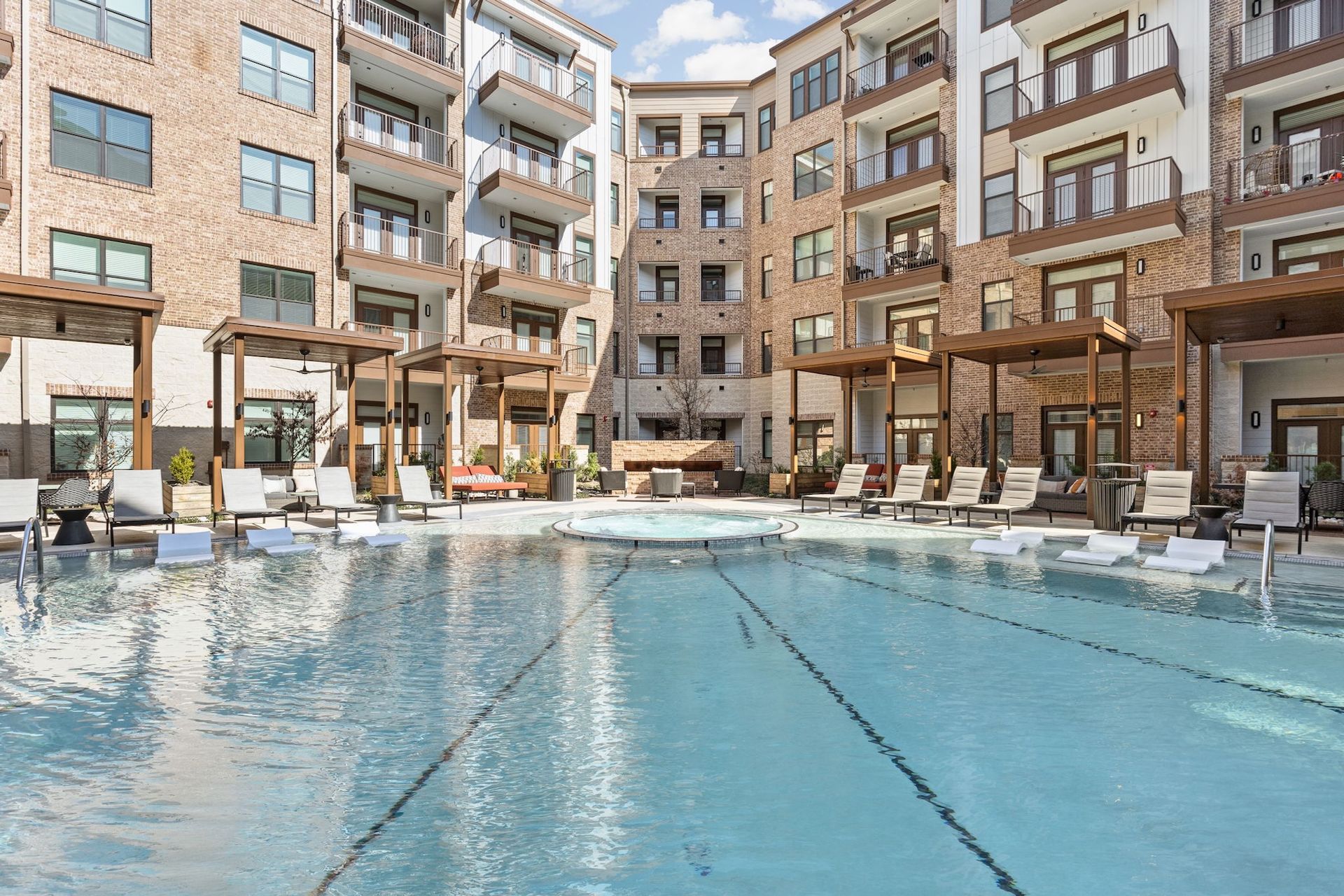 A swimming pool in a courtyard surrounded by multi-story brick apartment buildings with balconies and poolside cabanas.