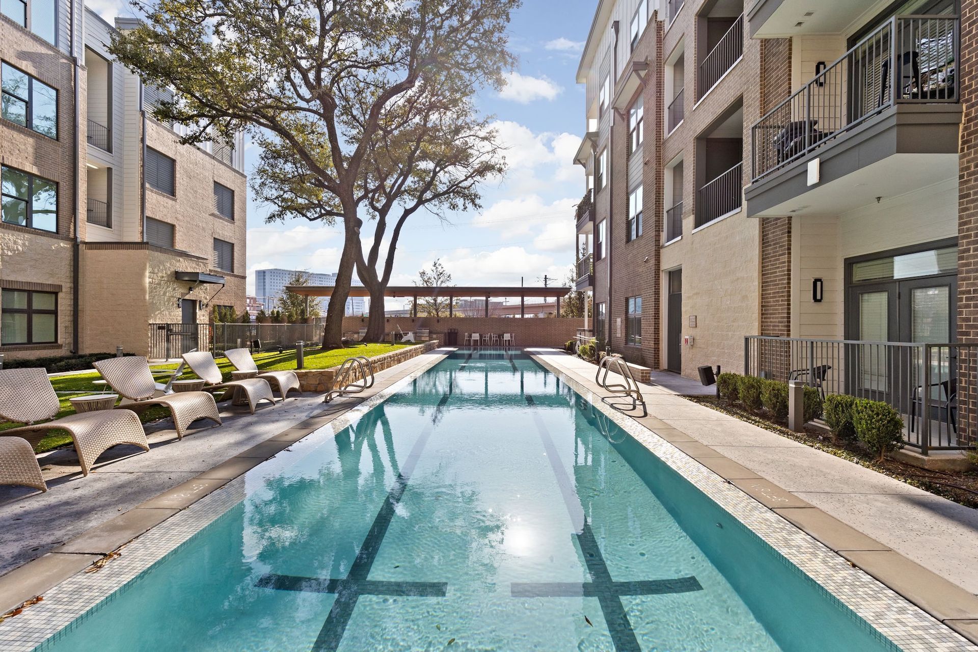 A narrow lap pool between two multi-story apartment buildings, with lounge chairs on the left and a sunny courtyard area.
