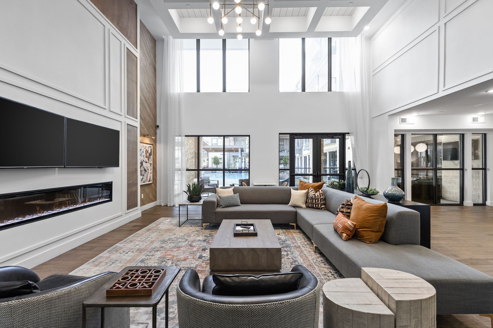 A spacious, modern lounge with a large gray sectional sofa, fireplace, patterned area rug, and high ceilings with windows.