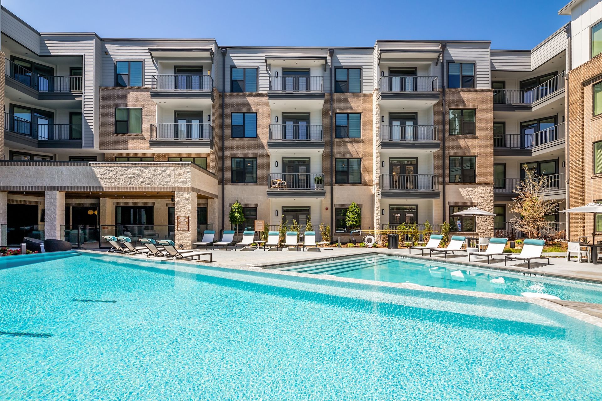 An outdoor resort-style swimming pool at a modern, multi-story apartment complex with lounge chairs on the patio.