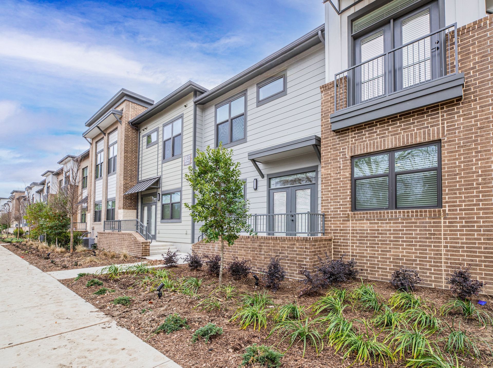 A row of multi-story townhomes with brick and light-colored siding, a sidewalk, and a landscaped bed in the foreground.