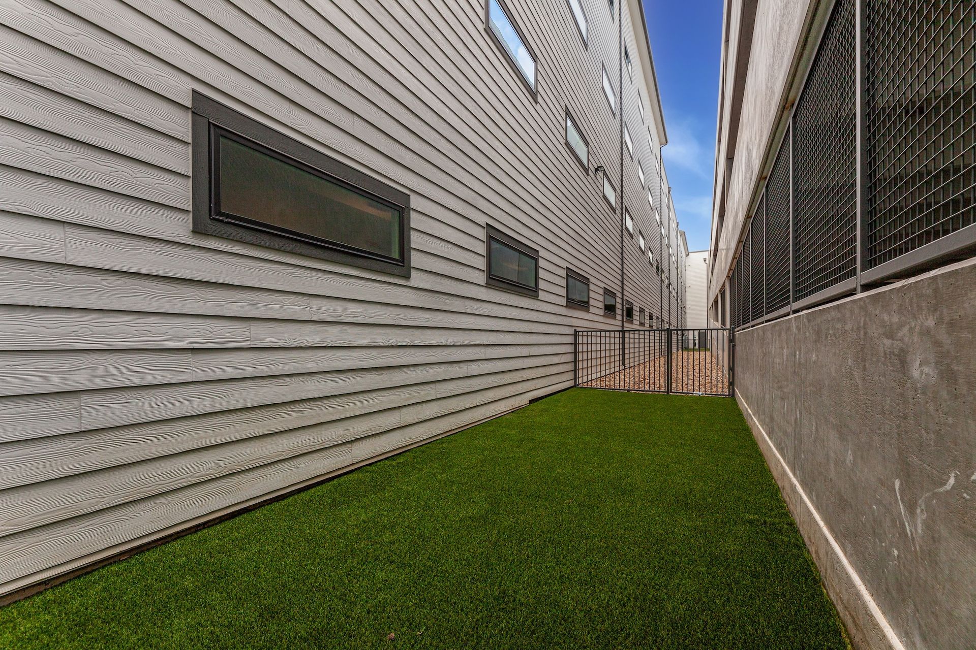 A narrow side yard with green artificial grass between two buildings, one with horizontal siding and small windows.