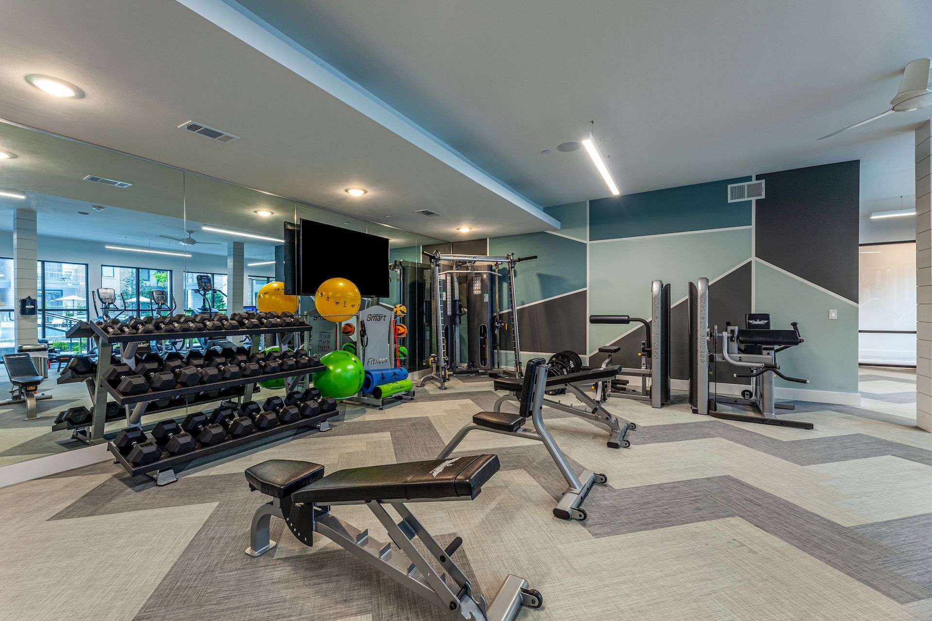 A brightly lit apartment fitness center featuring a weight rack, exercise benches, yoga balls, and strength machines.