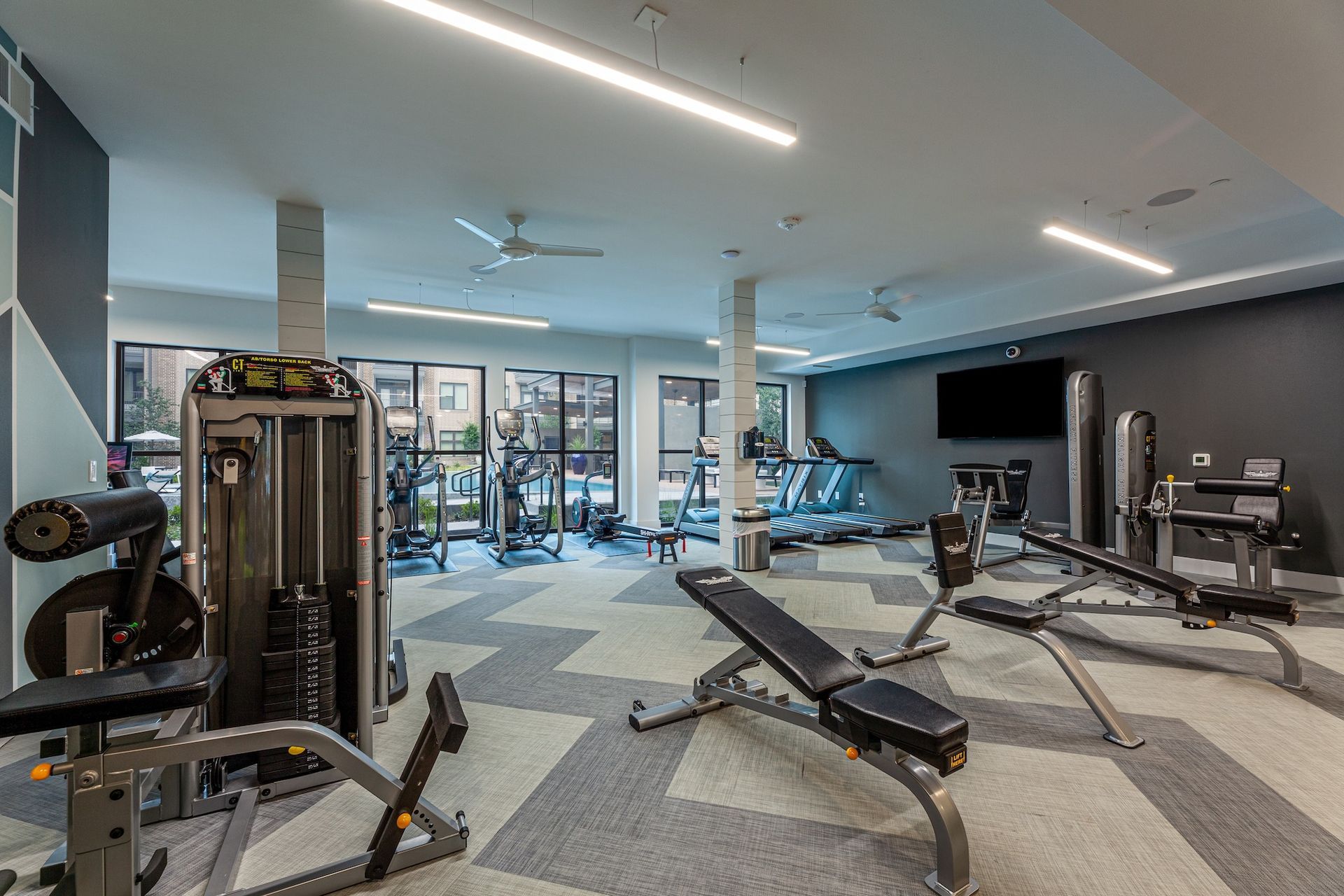 Modern fitness center with weight machines, benches, and cardio equipment on a gray patterned floor.