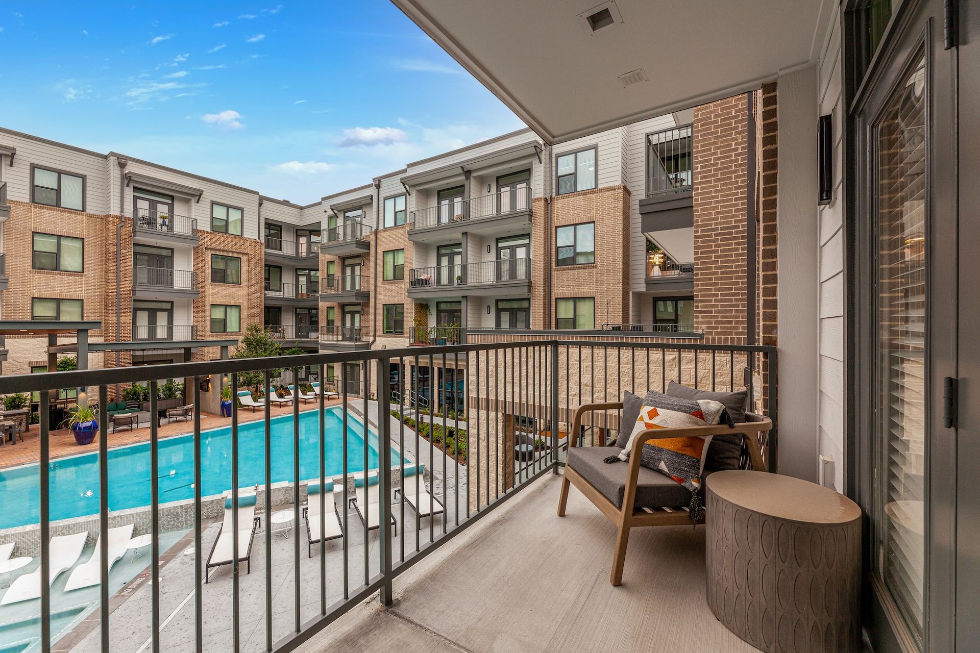 A balcony with a wooden chair and side table overlooking a luxury apartment complex pool area on a sunny day.