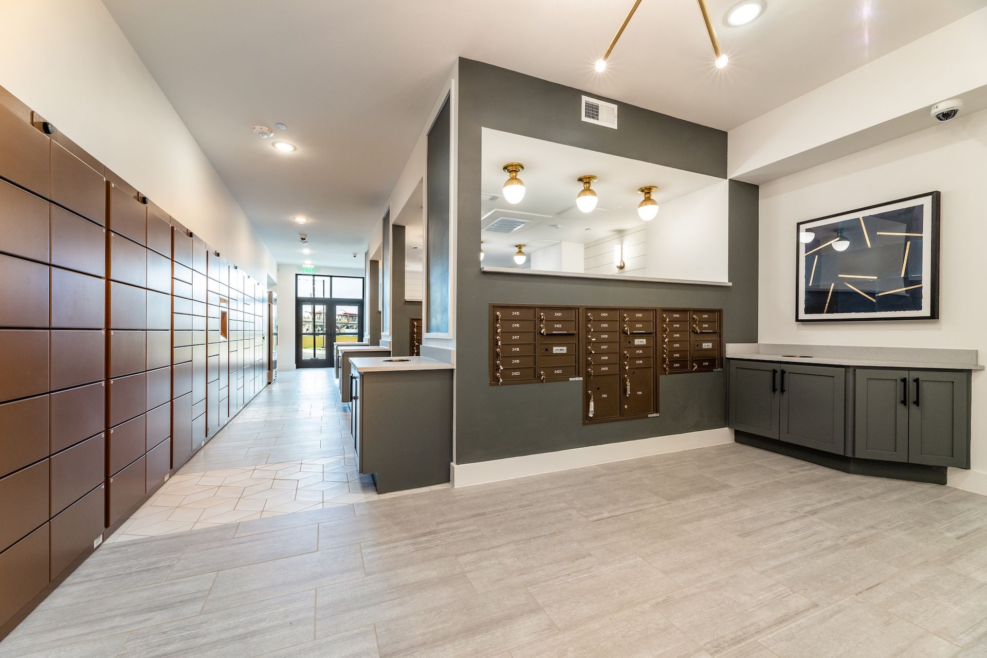 A well-lit residential mail and parcel room with tiled floors, dark metal mailboxes, and modern gray cabinetry.