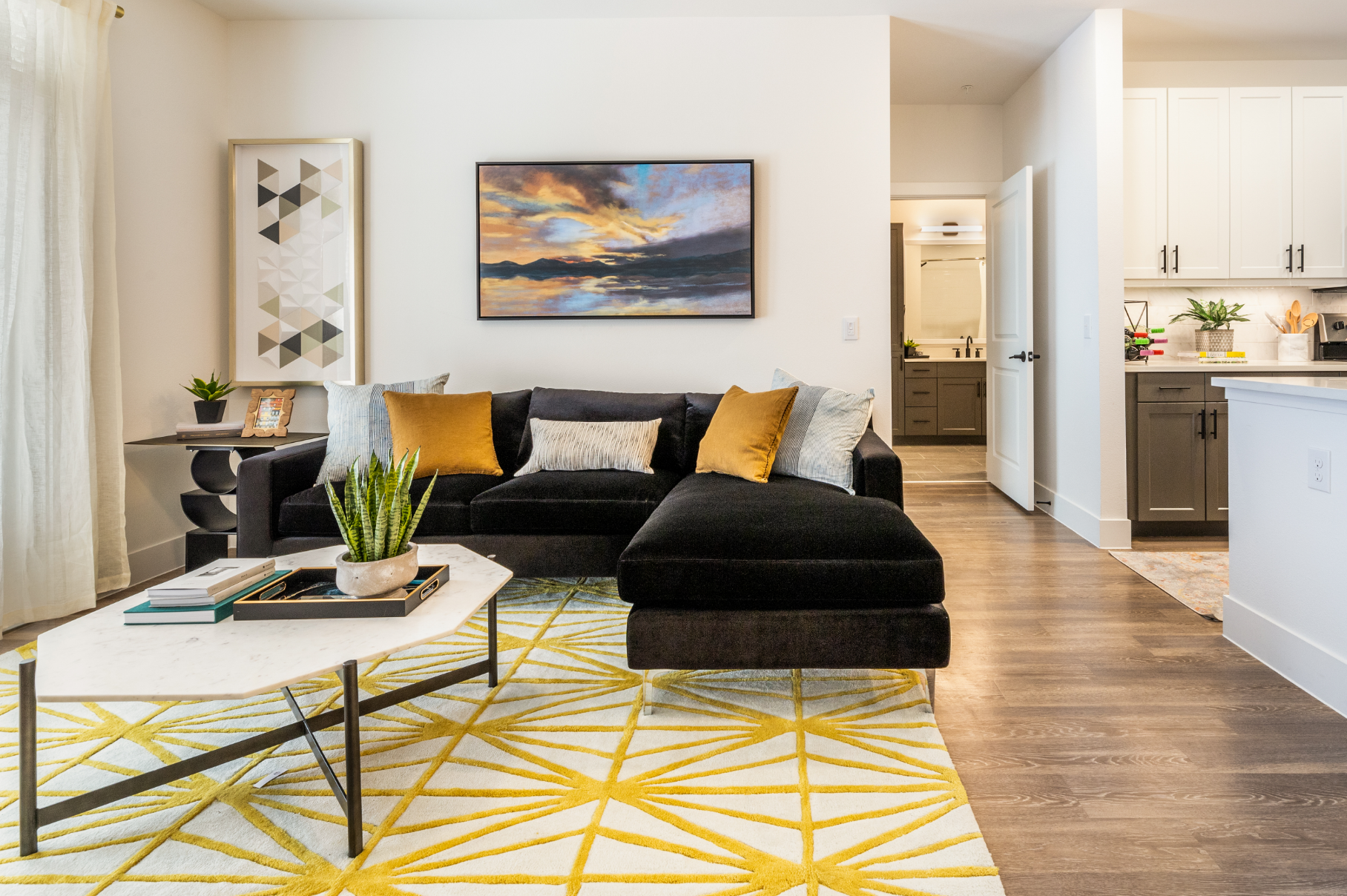 Living room with black sectional sofa, yellow rug, art, and open to kitchen and hallway.