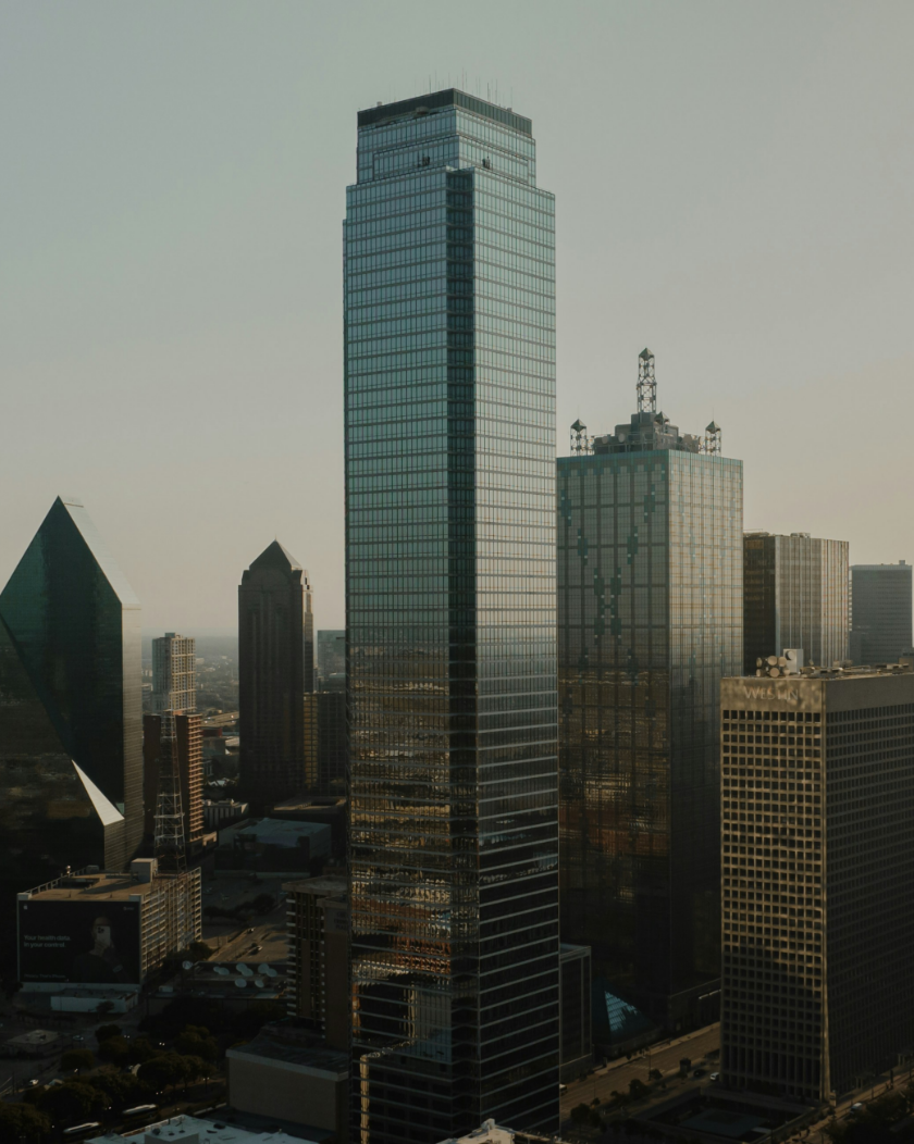 Tall glass skyscraper amidst a cityscape, reflecting sunlight. Other buildings in background, daytime.