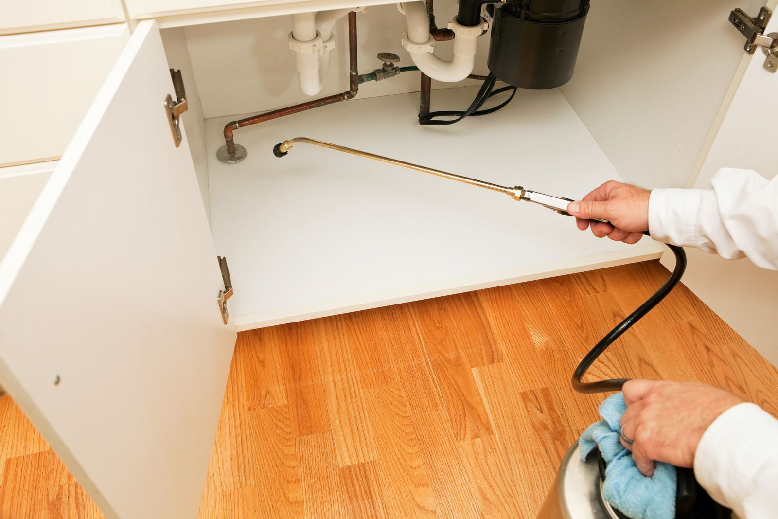 Person spraying under a kitchen sink with a pesticide, for pest control.