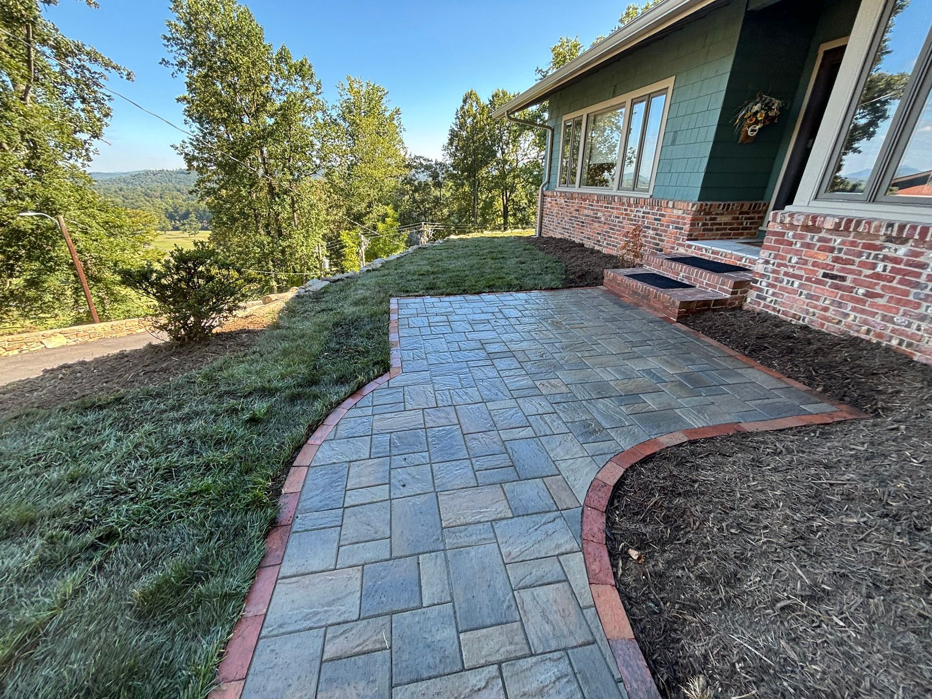 Paver walkway leading to a house with brick and green siding. The walkway has a red brick border.