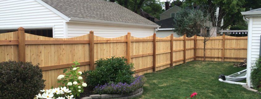 Wooden fence surrounds a backyard with green grass and flower beds. Houses are visible in the background.