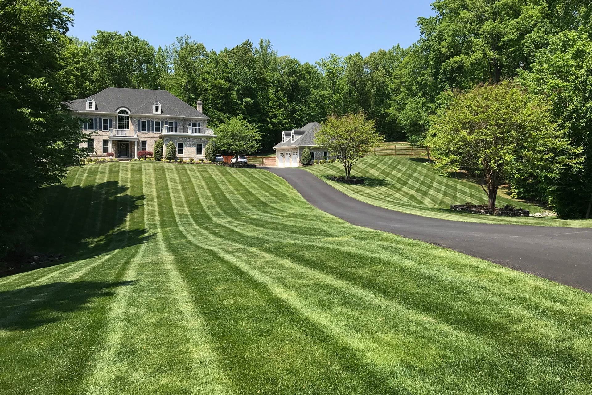 Large house on a hill with striped lawn and asphalt driveway, trees in the background, sunny day.