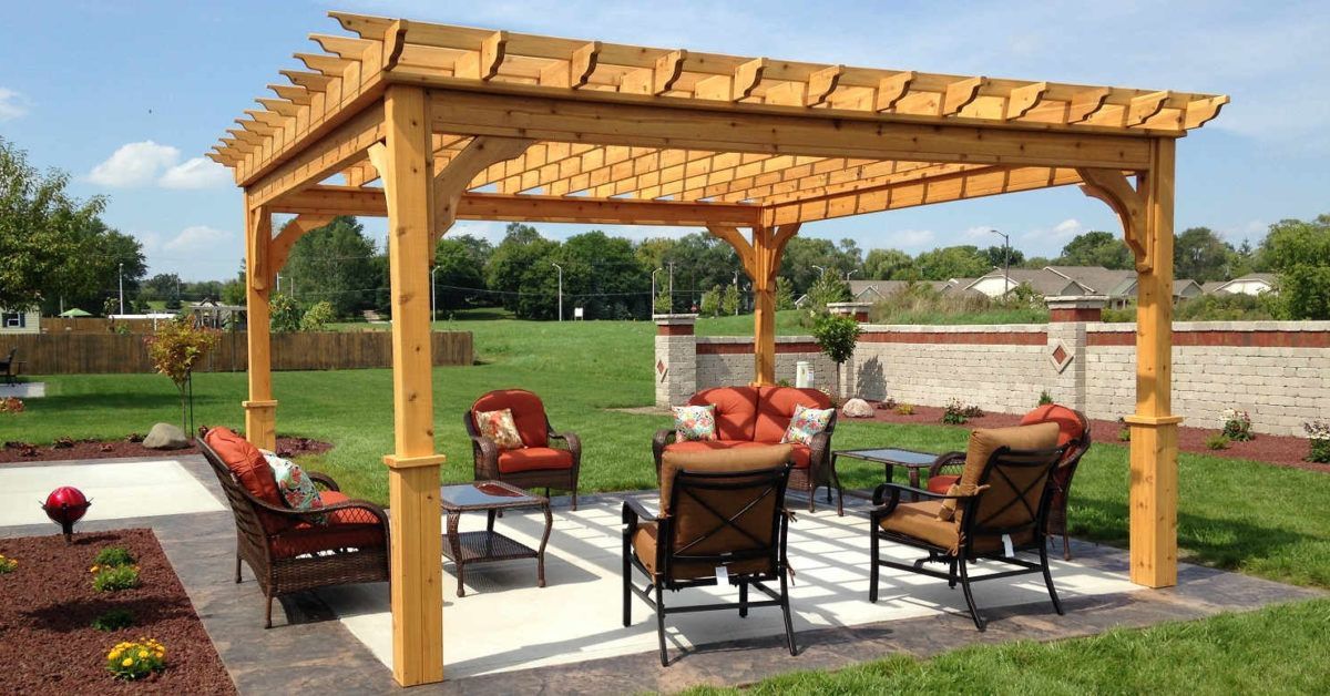 Wooden pergola over outdoor seating area: chairs, table, brick patio, green lawn, blue sky.