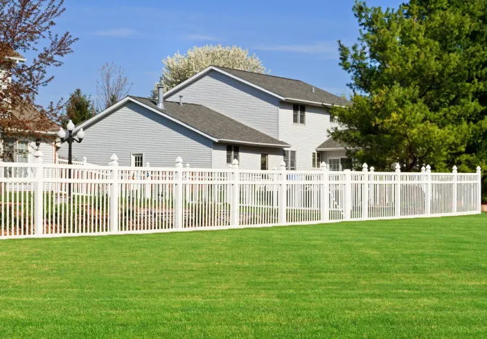 White picket fence encloses a green lawn and two-story gray house under a blue sky.