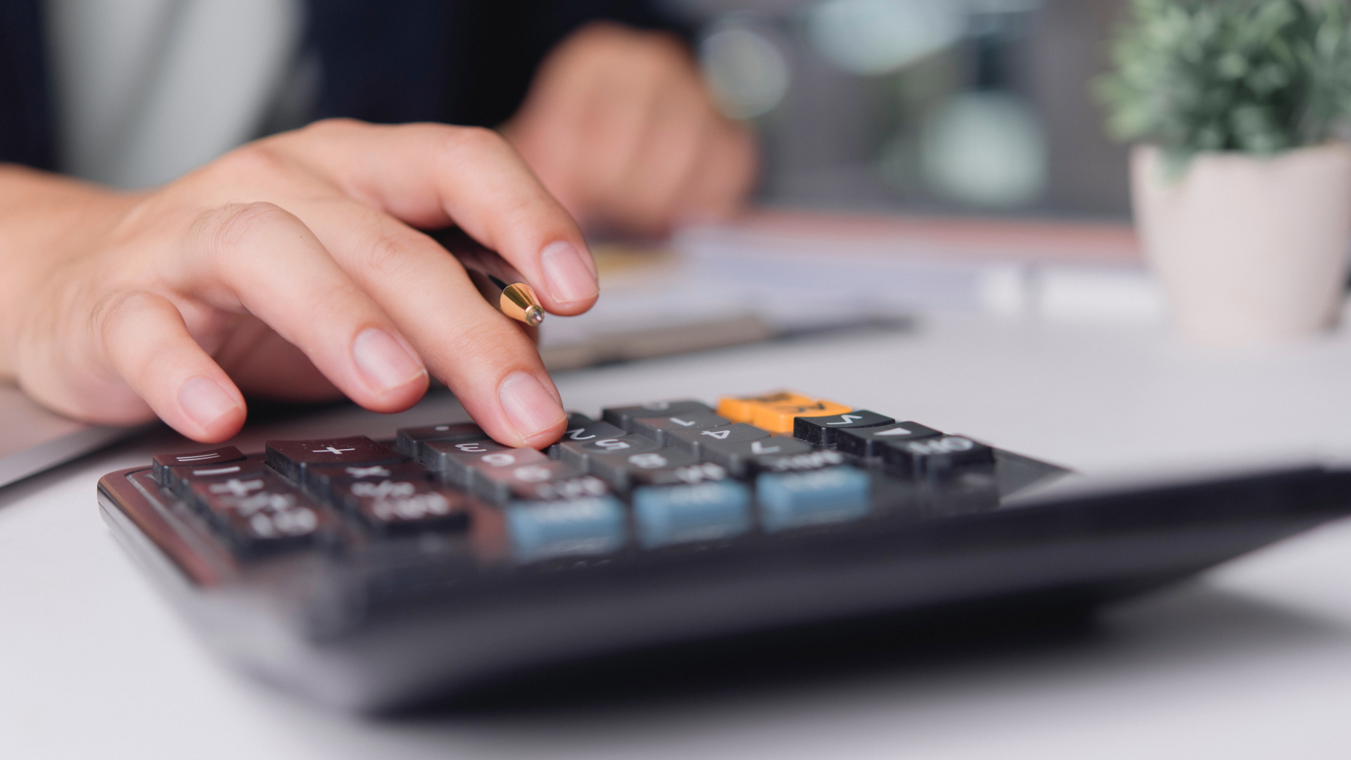 Person using a calculator with a notebook, pen, and stack of cash on a wooden surface.
